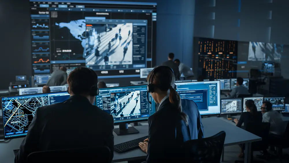 male and female sat in front of multiple monitors watching the footage from cameras on a street in a communications room setting
