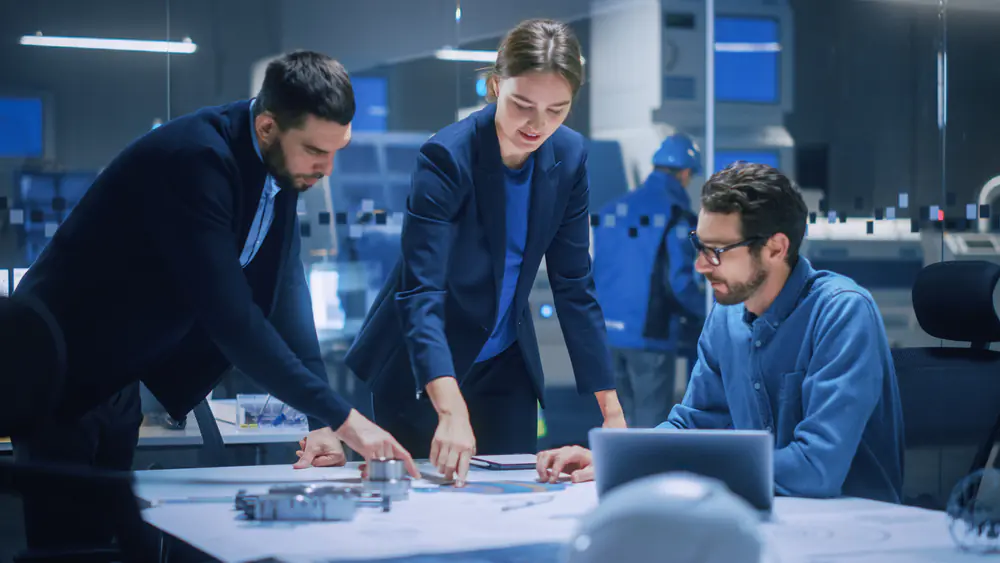 Two males and a female around a table pointing and looking at documents