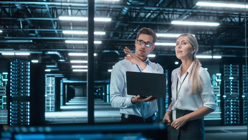 A male and female looking into the distance engaging in conversation whilst holding a tablet device in a data centre