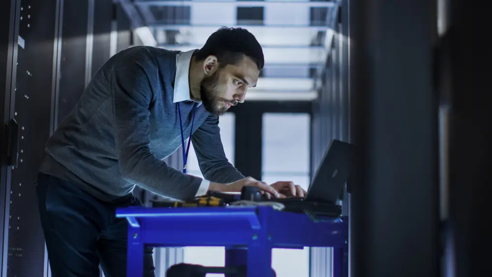 Male IT technician hunched over at a laptop working in front of a data centre rack